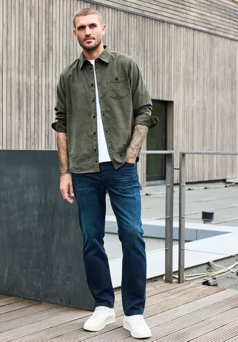 Mann in grüner Cord-Hemdjacke, weißem Shirt, blauen Jeans und weißen Sneakern auf Dachterrasse.