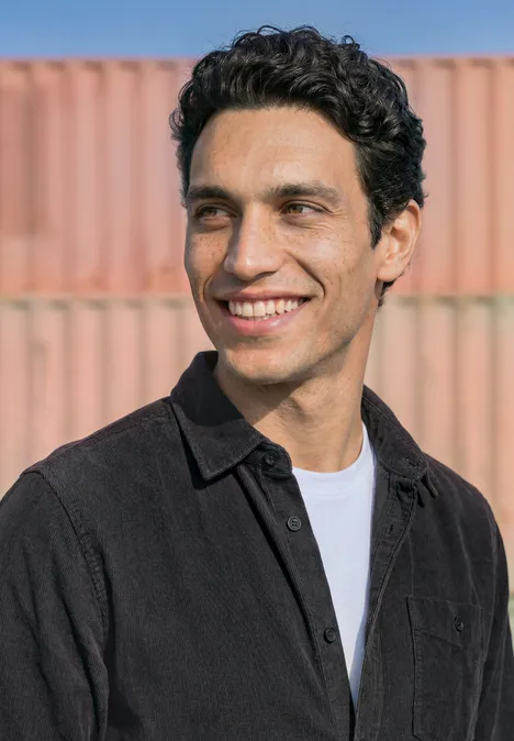 Ein lächelnder Mann mit dunklen Locken, weißem Shirt und schwarzer Jacke vor rosa-oranger Containerwand.