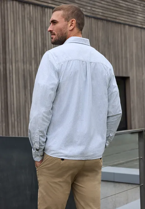 Ein Mann in hellem Hemd und beigen Hosen lehnt entspannt an einem Geländer vor einer Holzfassade.