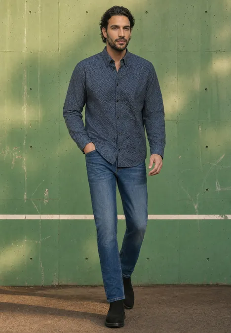 Een man in een donkerblauw hemd en jeans staat ontspannen voor een groene muur