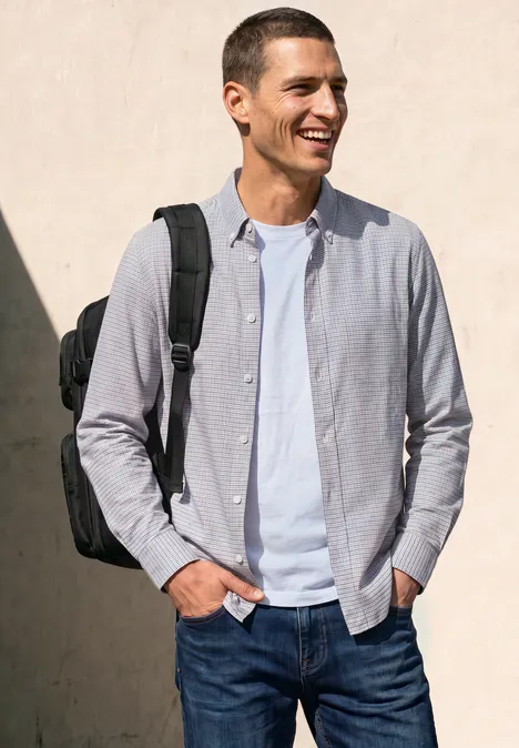 Een lachende man in jeans, wit T-shirt en lichtgrijs overhemd met zwarte rugzak voor een lichte muur.