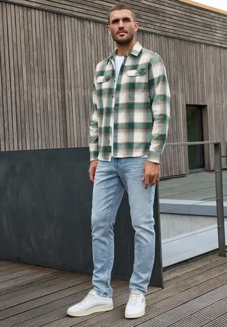 Ein Mann steht auf einer Terrasse vor Holzfassade, trägt grün-weiß kariertes Hemd, helle Jeans und weiße Sneaker.