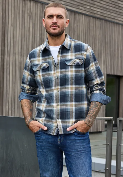 Ein bärtiger Mann in karierter Flanelljacke und Jeans lehnt an einem Geländer vor einer Holzfassade.