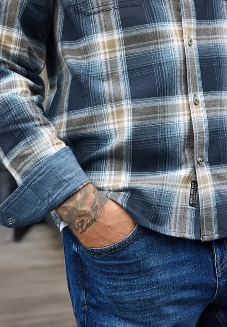 Ein Mann trägt ein blau kariertes Hemd, steckt die Hand in die Jeans und zeigt ein Tattoo am Handgelenk.