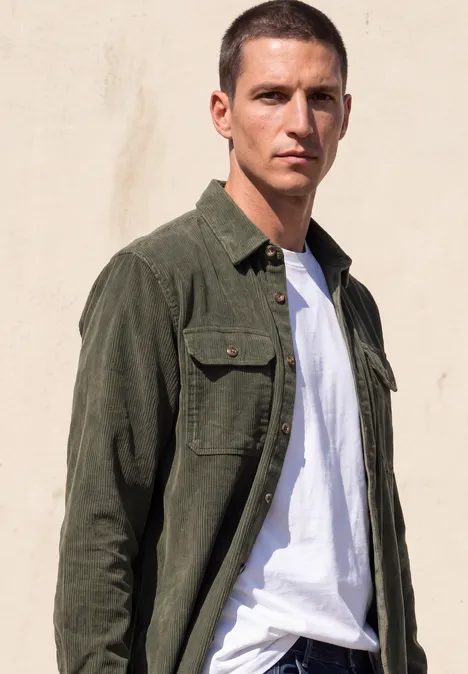 Un homme aux cheveux courts porte un t-shirt blanc et une chemise kaki ouverte devant un mur clair.