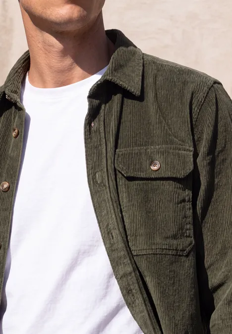 Un homme porte une veste en velours côtelé olive sur un t-shirt blanc devant un mur clair.