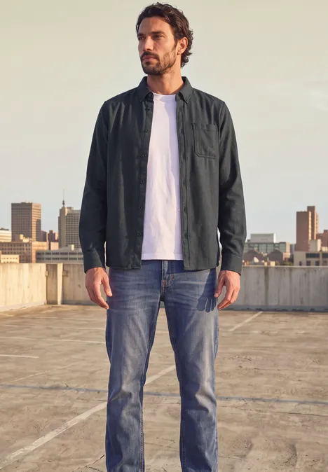 Ein Mann in Jacke, weißem T-Shirt und Jeans steht auf einem Parkdach vor Stadtkulisse.
