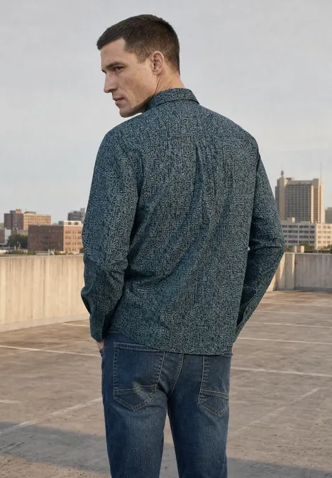 Ein Mann in Jeans und gemusterter Jacke steht auf einem Dach mit Stadtsilhouette im Hintergrund.