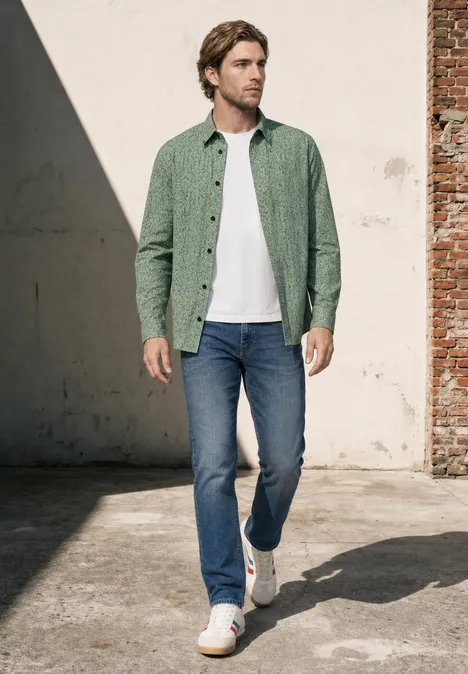 Ein Mann in grünem Hemd, weißem T-Shirt, Jeans und weißen Sneakern geht entspannt an einer Backsteinwand vorbei.