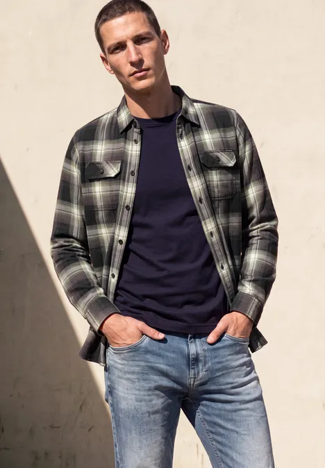 Mann vor heller Wand, trägt dunkles T-Shirt, kariertes Holzfällerhemd und Jeans.