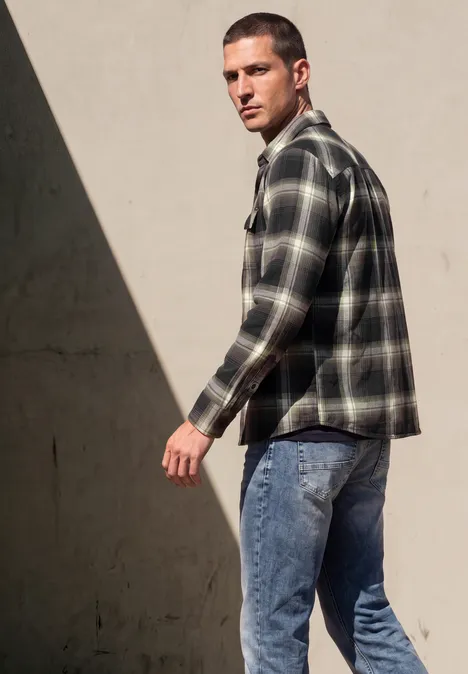 Ein Mann in kariertem Hemd und Jeans läuft vorbei und schaut über die Schulter vor einer hellen Wand.
