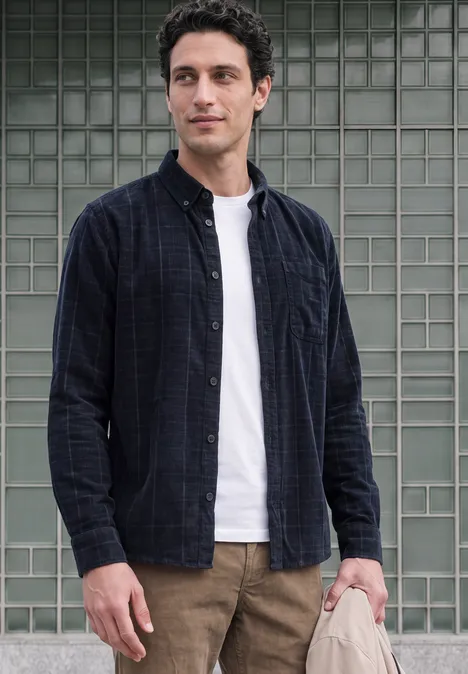 Un jeune homme se tient devant un mur de carreaux de verre, vêtu d’un t-shirt blanc, d’une chemise à carreaux ouverte et d’un pantalon marron.