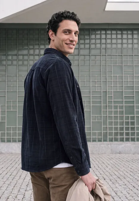 Un jeune homme en jean et chemise sombre regarde par-dessus l’épaule devant un mur à grille.