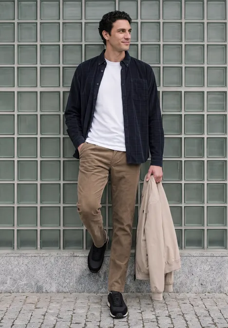 Un homme marche devant un mur de verre, porte pantalon beige, t-shirt blanc, veste sombre et tient un manteau.