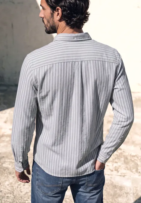 Ein Mann im hellblauen, gestreiften Hemd und Jeans steht mit dem Rücken zur Kamera vor einer grauen Wand.