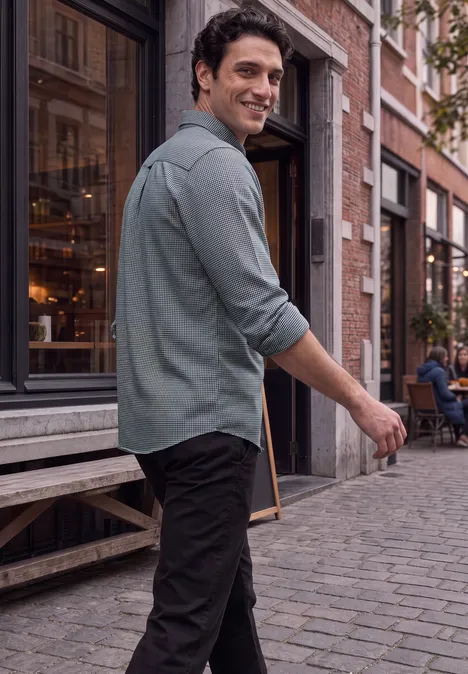 Un homme en chemise verte et pantalon noir marche en souriant sur une rue pavée devant un café.