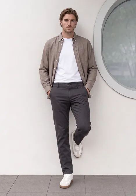 Un homme est appuyé contre un mur, vêtu d’un t-shirt blanc, d’un pantalon gris, d’une veste brune et de baskets blanches.