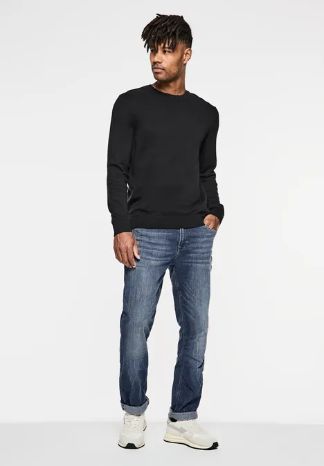 Jean used coupe droite indigo blue wash Mann mit schwarzem Pullover, Jeans und weißen Sneakers steht und blickt zur Seite.