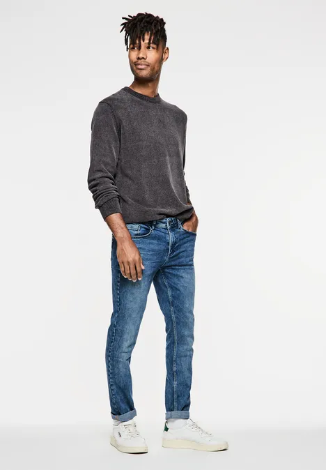 Mann mit dunklem Pullover, blauer Jeans und weißen Sneakers steht locker.