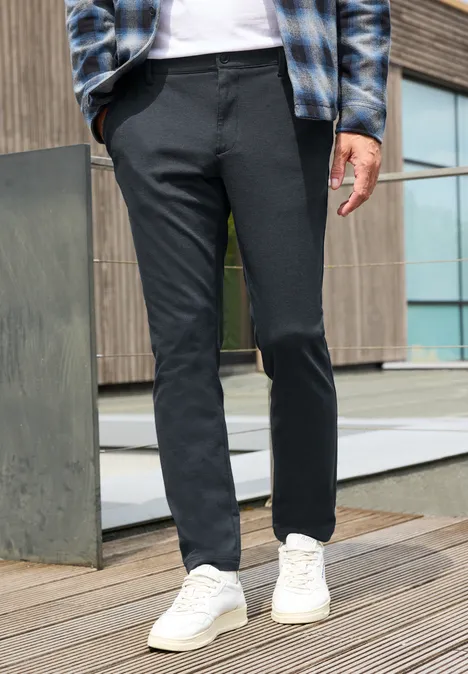 Ein Mann trägt dunkelgraue, schmale Hose und weiße Sneaker auf einer Holzterrasse vor einem modernen Gebäude.