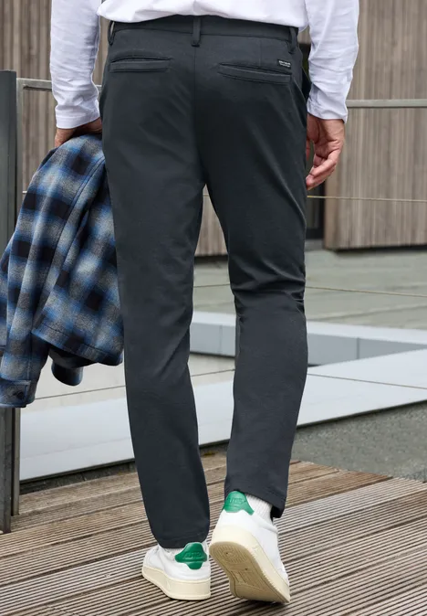 Eine Person steht auf einer Holzplattform, trägt schwarze Hose und weißes Hemd, hält ein kariertes Jackett.