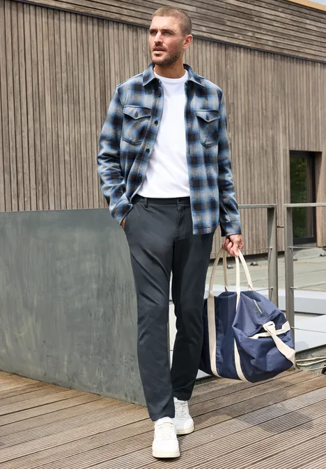 Ein Mann in karierter Hemdjacke, weißem T-Shirt, grauer Hose und weißen Sneakern trägt eine Reisetasche.