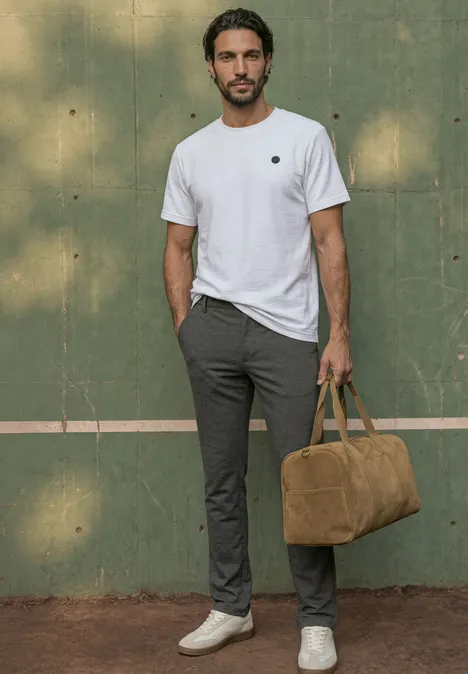 Ein Mann mit weißen Sneakers, grauen Hosen und weißem T-Shirt hält eine braune Tasche vor grünem Hintergrund.