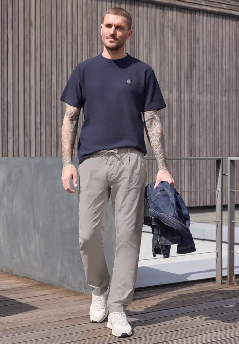 Mann mit Tattoos geht auf Holzpodest, trägt blaues T-Shirt, graue Hose, weiße Sneaker und hält Jacke.