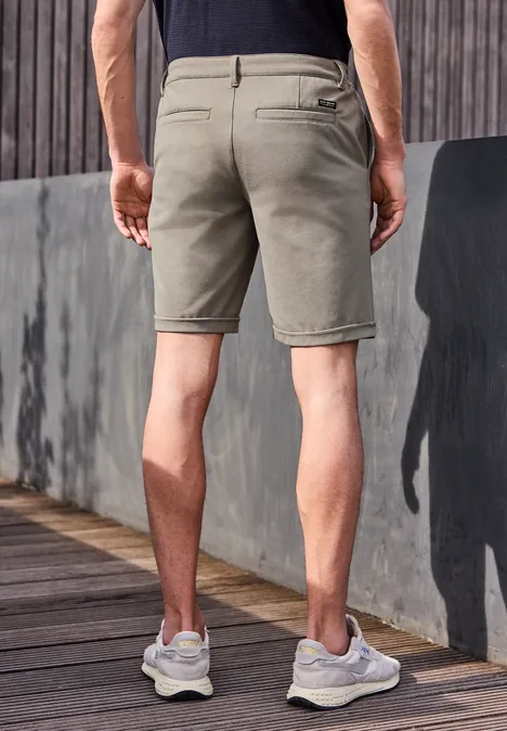 Rückenansicht eines Mannes in khakifarbenen Shorts und weißen Sneakern auf Holzterrasse vor grauer Wand.