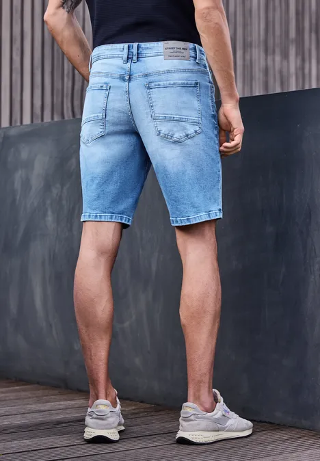 Homme de dos en short en jean bleu clair, adossé à un mur mixte bois et béton.