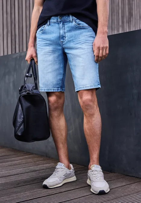 Homme en short en jean et baskets, tenant un sac noir, debout devant un mur.
