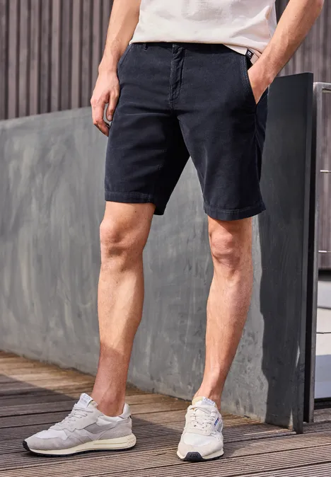 Mann in weißem T-Shirt, schwarzen Chino-Shorts und weißen Sneakern steht auf Holzdeck vor moderner Wand.