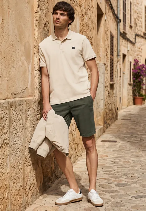 Mann in beigem Poloshirt, grünen Shorts und weißen Sneakern hält Jacke und steht in sonniger Altstadtgasse.
