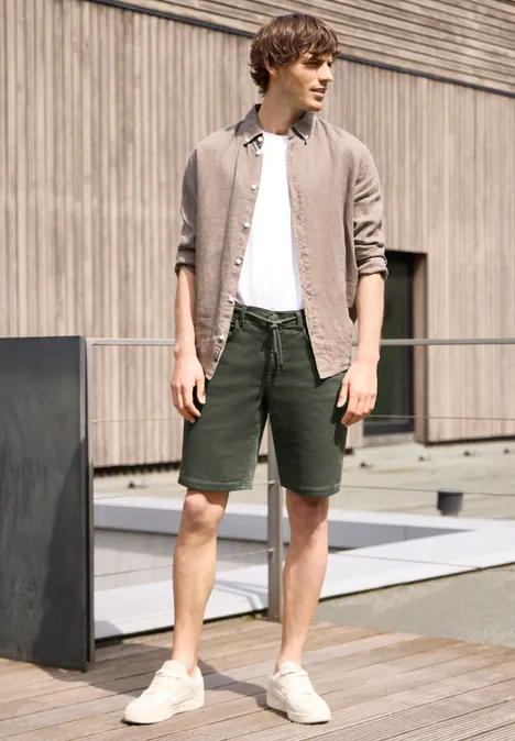 Man in wit T-shirt, beige overhemd open, groene korte broek en witte sneakers voor houten gevel.