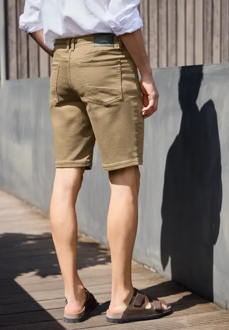 Een man in beige short, wit overhemd en sandalen staat zijwaarts voor een houten wand in zonlicht.