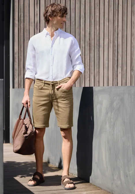 Een man in een wit overhemd en bruine korte broek met tas, staand voor een houten gevel.