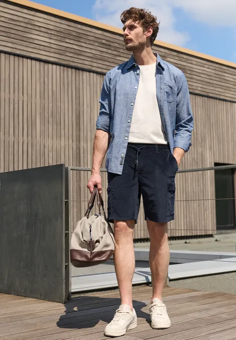 Een man in overhemd, t-shirt, korte broek en sneakers staat buiten en houdt een reistas vast.