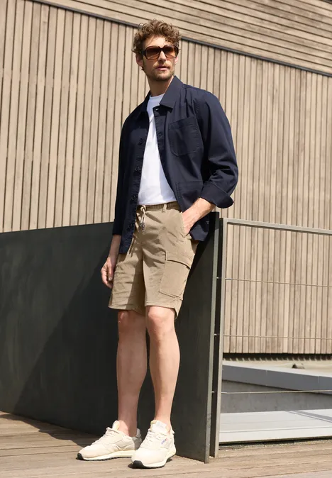 Ein Mann in beigen Shorts, weißem T-Shirt und dunkelblauer Hemdjacke lehnt an einer Holzfassade.