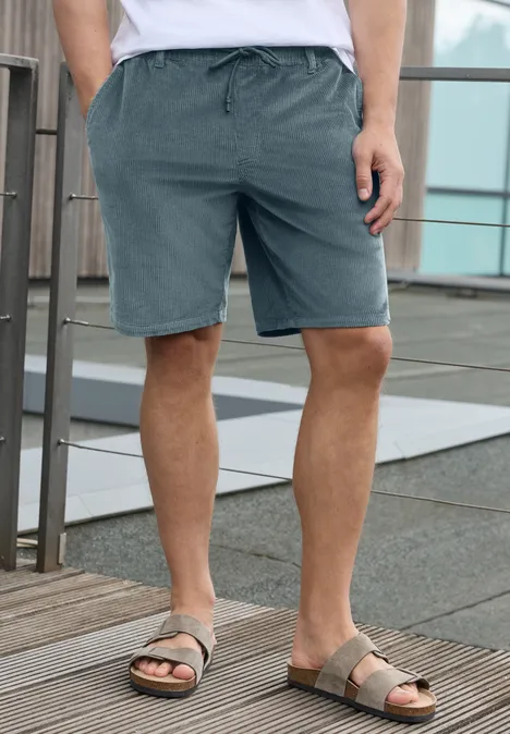 Mann steht auf Holzterrasse in grauen Shorts und Sandalen vor Glas- und Metallgeländer.