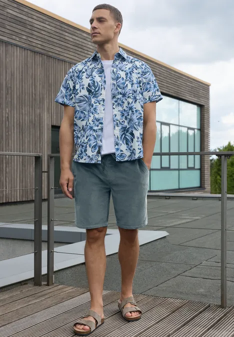 Man op dakterras voor hout-glasgebouw, draagt blauw gebloemd overhemd, wit shirt, grijze shorts en sandalen.