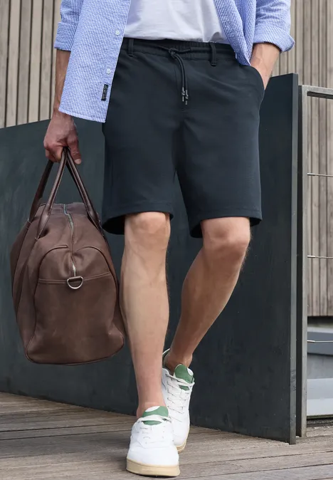 Jogg Chino Shorts aus Jersey mit Flexbund dark ink blue Ein Mann in schwarzen Shorts und weißen Sneakern hält eine braune Lederreisetasche und geht.