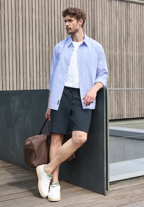 Jogg Chino Shorts aus Jersey mit Flexbund dark ink blue Junger Mann in lässigem Outfit mit Shorts, Hemd, Sneakern und Leder-Reisetasche vor urbaner Kulisse
