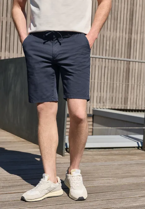Ein Mann trägt dunkelblaue Shorts und weiße Sneaker und steht auf einer Holzterrasse.