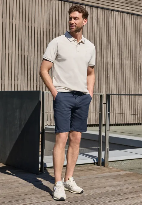 Ein Mann in Polohemd und Shorts lehnt entspannt an einem Geländer auf einer Holzterrasse.