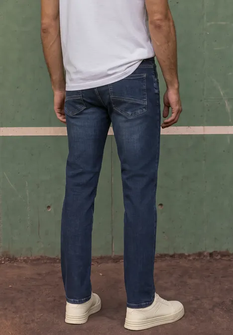 Un homme vu de dos, en jean bleu et baskets blanches, devant un mur vert.