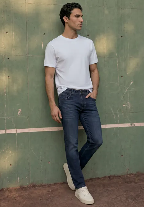 Un homme en t-shirt blanc et jean bleu appuyé contre un mur vert.