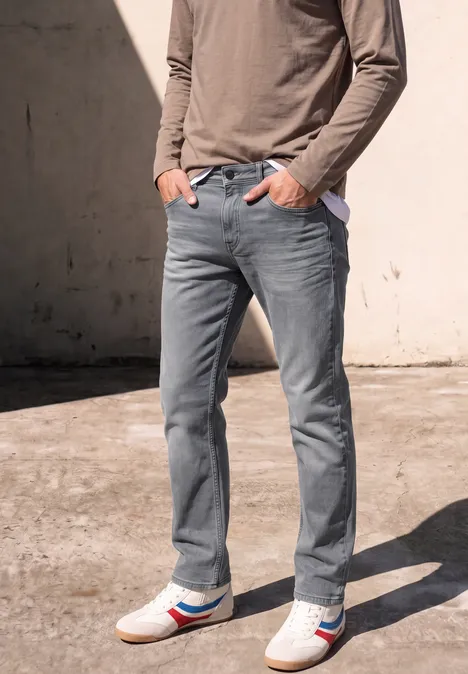 Homme en jean gris, haut à manches longues et baskets blanches à rayures rouge et bleu devant un mur.