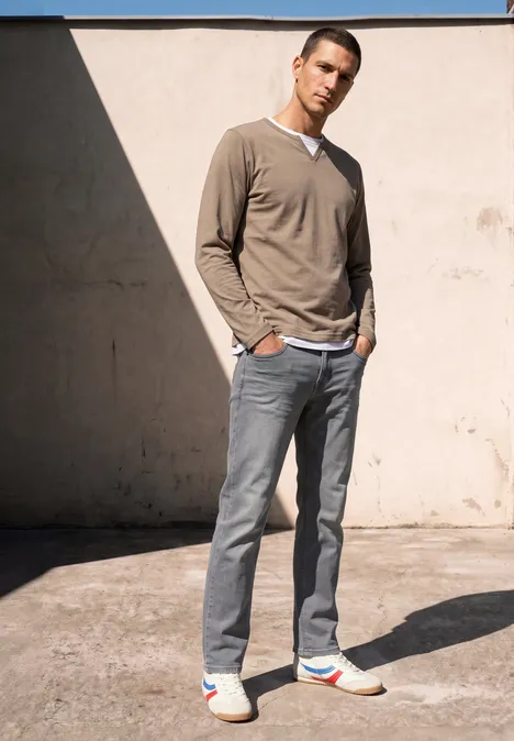Un homme sportif en pull beige, jean gris et baskets colorées se tient détendu devant un mur ensoleillé.