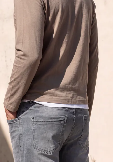 Homme vu de dos portant un jean gris et un tee-shirt à manches longues beige, devant un mur clair.