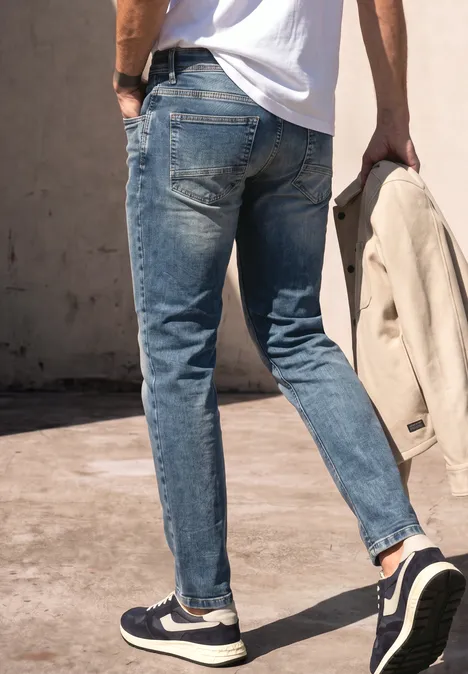 Ein Mann in blauen Jeans und Sneakern hält eine beige Jacke und geht über einen Gehweg.